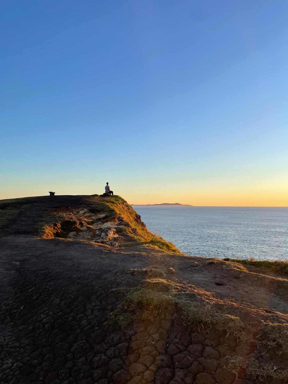 A solitary figure seated on a coastal cliff at sunset